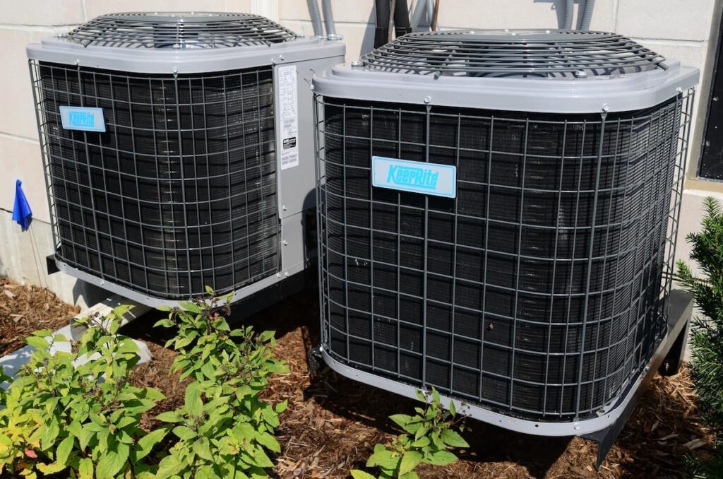 Two Keeprite outdoor air conditioning units are installed side by side on a concrete pad next to a building, surrounded by mulch and small plants.